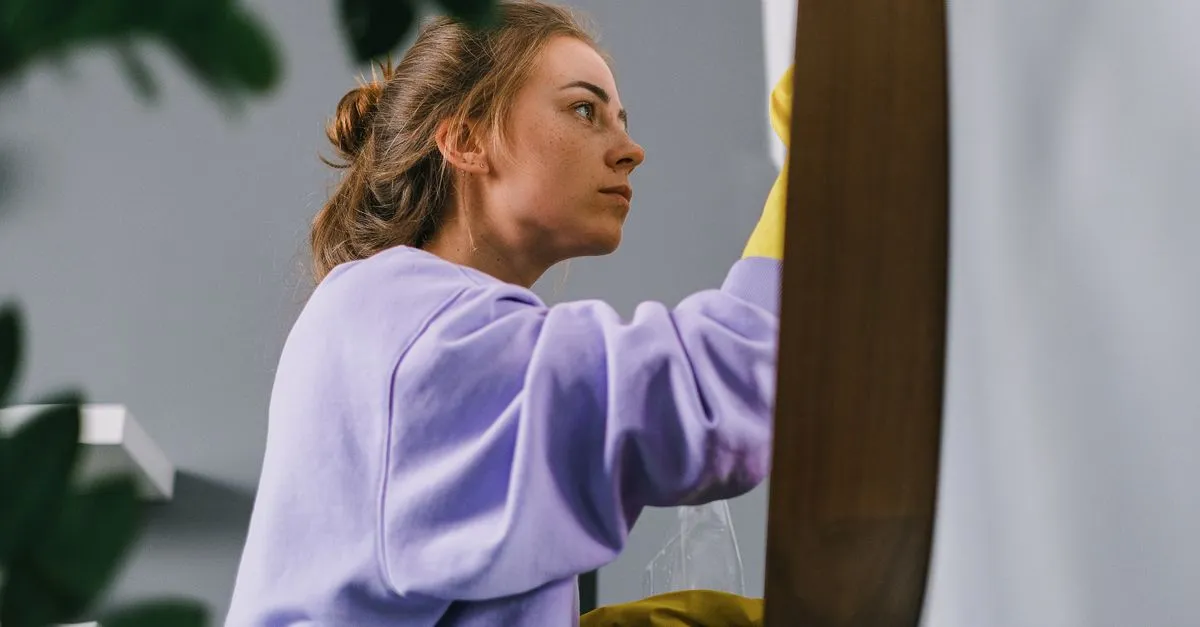 Side view of young focused female in latex gloves standing and cleaning mirror in light room in daytime